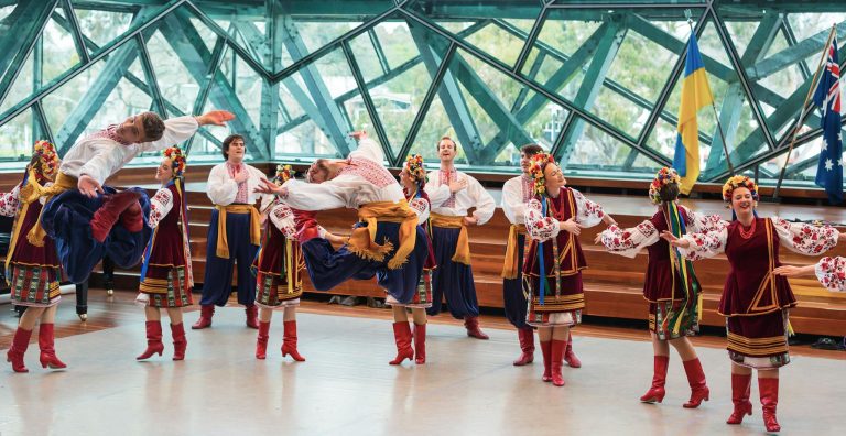 Group performing vibrant traditional folk dance indoors with flags visible.