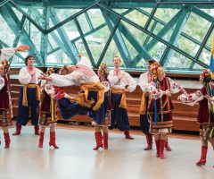 Group performing vibrant traditional folk dance indoors with flags visible.