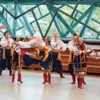 Group performing vibrant traditional folk dance indoors with flags visible.