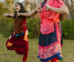 Two women in colorful clothing dancing on grass with hand gestures and smiles.
