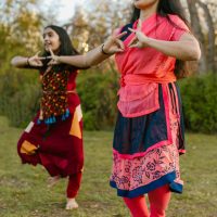 Two women in colorful clothing dancing on grass with hand gestures and smiles.